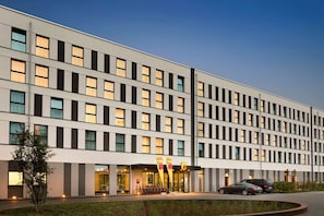 A modern hotel building with a row of uniformly sized windows, a visible entrance with a sign, and a parking area with cars and bicycles. - Freiburg (Freiburg im Breisgau)