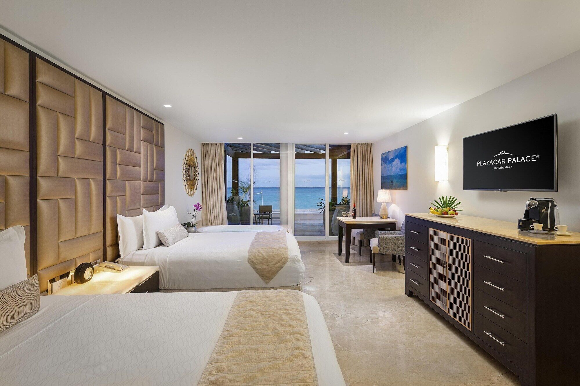 A hotel room with two beds, a desk, a TV, and a view of the sea.