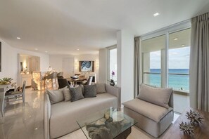 Una sala de estar moderna con sofá, sillón y mesa de centro, un comedor con sillas y una mesa, y un ventanal con vistas al océano.