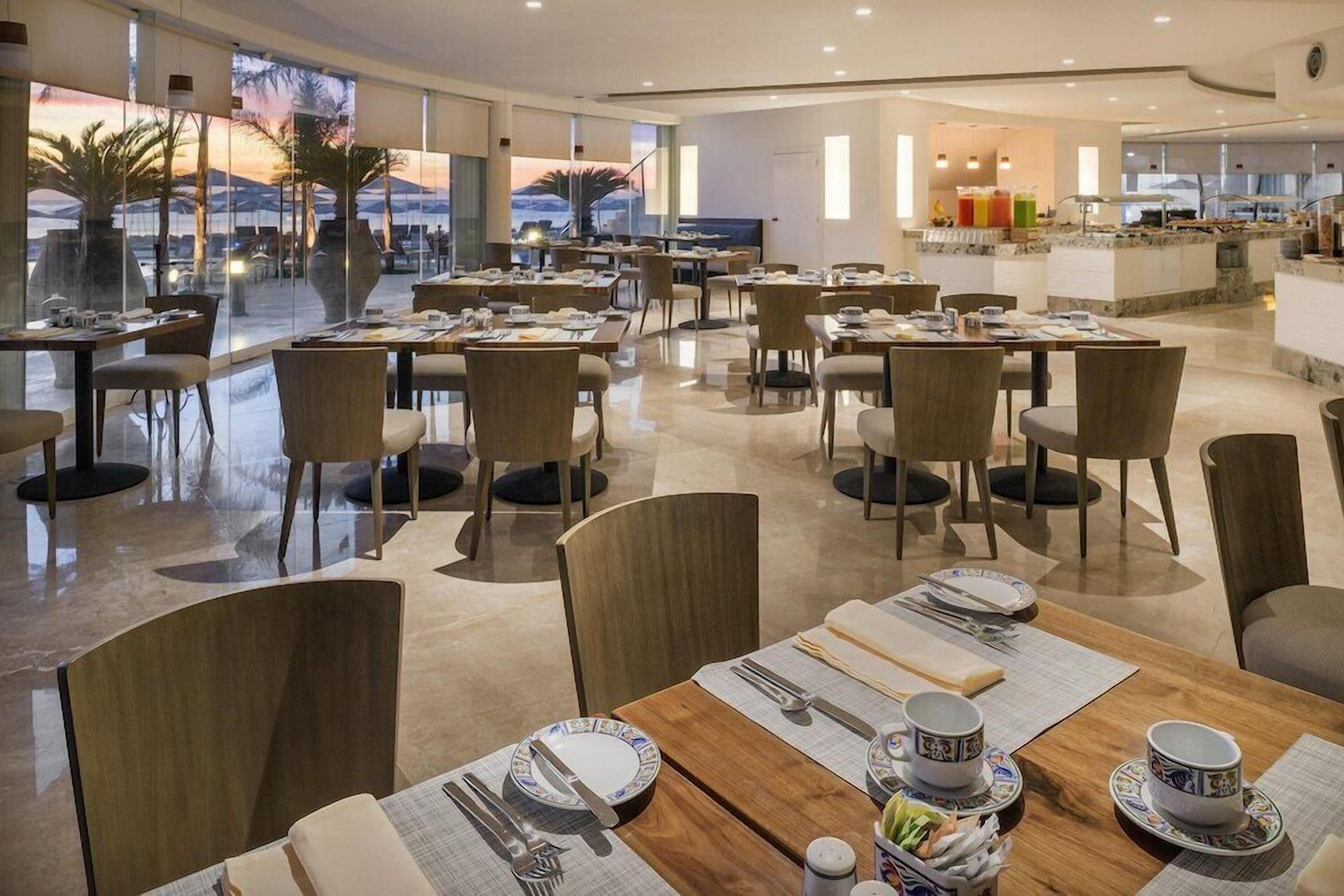 Een moderne eetruimte met gedekte tafels, een keuken op de achtergrond en uitzicht op palmbomen en bergen door grote ramen.