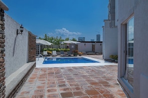 A swimming pool with lounge chairs, a brick patio, and a city skyline in the background. - Suites Mexico Plaza Campestre (León)