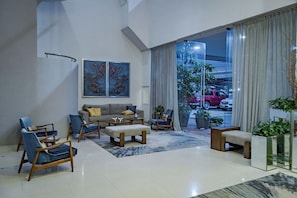 Un salon moderne avec un canapé, des fauteuils et une table basse. De grandes fenêtres sont présentes, ornées de rideaux, et on peut apercevoir un camion rouge à l’extérieur.