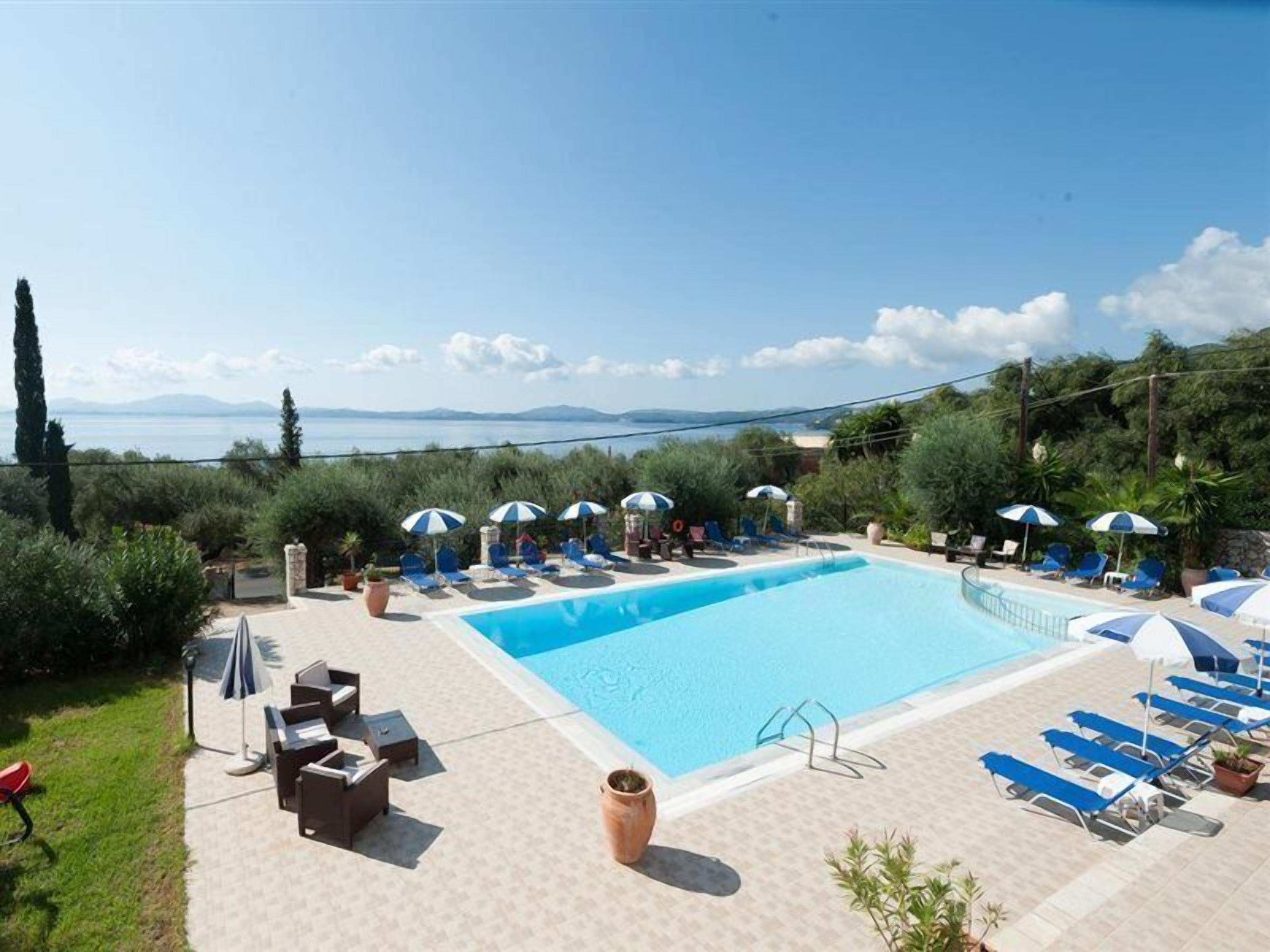 A pool area with lounge chairs and umbrellas, surrounded by greenery and a view of the sea.