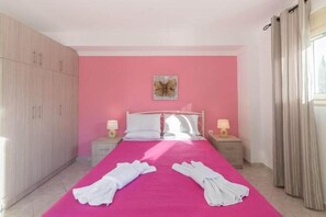 A bedroom with a pink wall, a bed with white linens, two bedside tables with lamps, and a wardrobe. - Crystal Blue Aparthotel (Corfu)