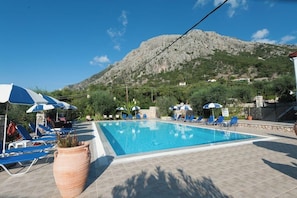 A swimming pool with lounge chairs and umbrellas, surrounded by greenery and a mountain in the background. - Crystal Blue Aparthotel (Corfu)