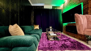 A living room with a green sofa, a pink armchair, a glass coffee table, and a large flat-screen TV.