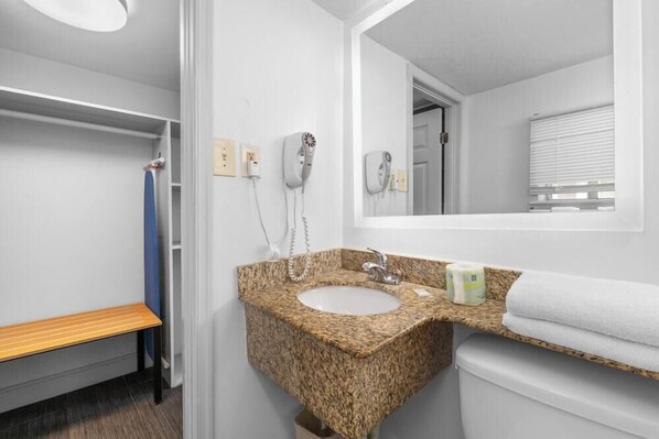 A bathroom with a granite sink, a mirror, a bench, and a toilet. - Travelodge by Wyndham Virginia Beach (Virginia Beach)