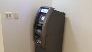 Kulmassa sijaitseva huone, jossa on harmaa pankkiautomaatti, pistorasia ja seinäpistoke.