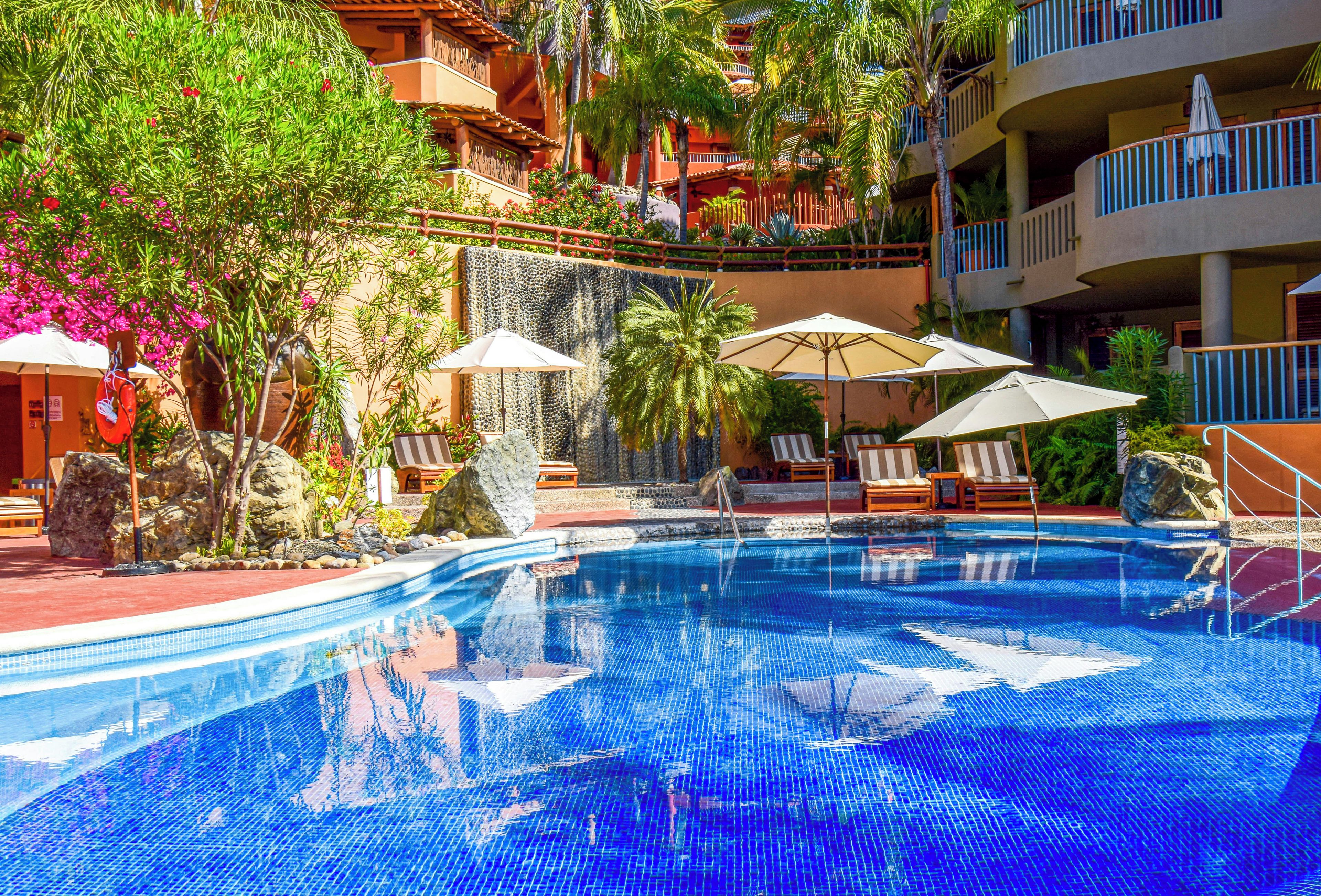 A swimming pool with lounge chairs and umbrellas, surrounded by tropical plants and a multi-story building with balconies.