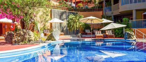 A swimming pool with lounge chairs and umbrellas, surrounded by tropical plants and a multi-story building with balconies.