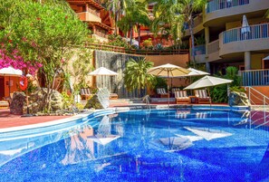 Ein Swimmingpool mit Liegestühlen und Sonnenschirmen, umgeben von tropischer Vegetation und einem mehrstöckigen Gebäude mit Balkonen.