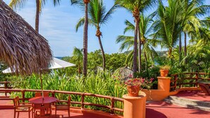 A tropical outdoor seating area with a thatched roof, palm trees, and a red-tiled floor. - Hilton Grand Vacations Club Zihuatanejo (Zihuatanejo)