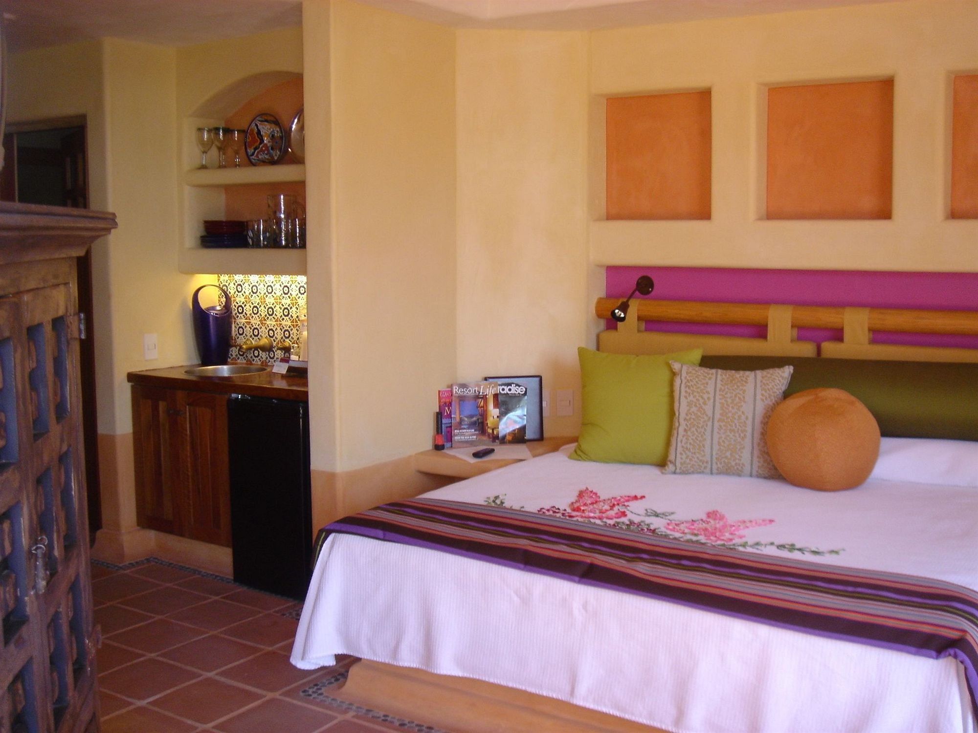 A hotel room with a bed, a small desk, a refrigerator, a shelf with decorative items, and a wall with a framed picture.