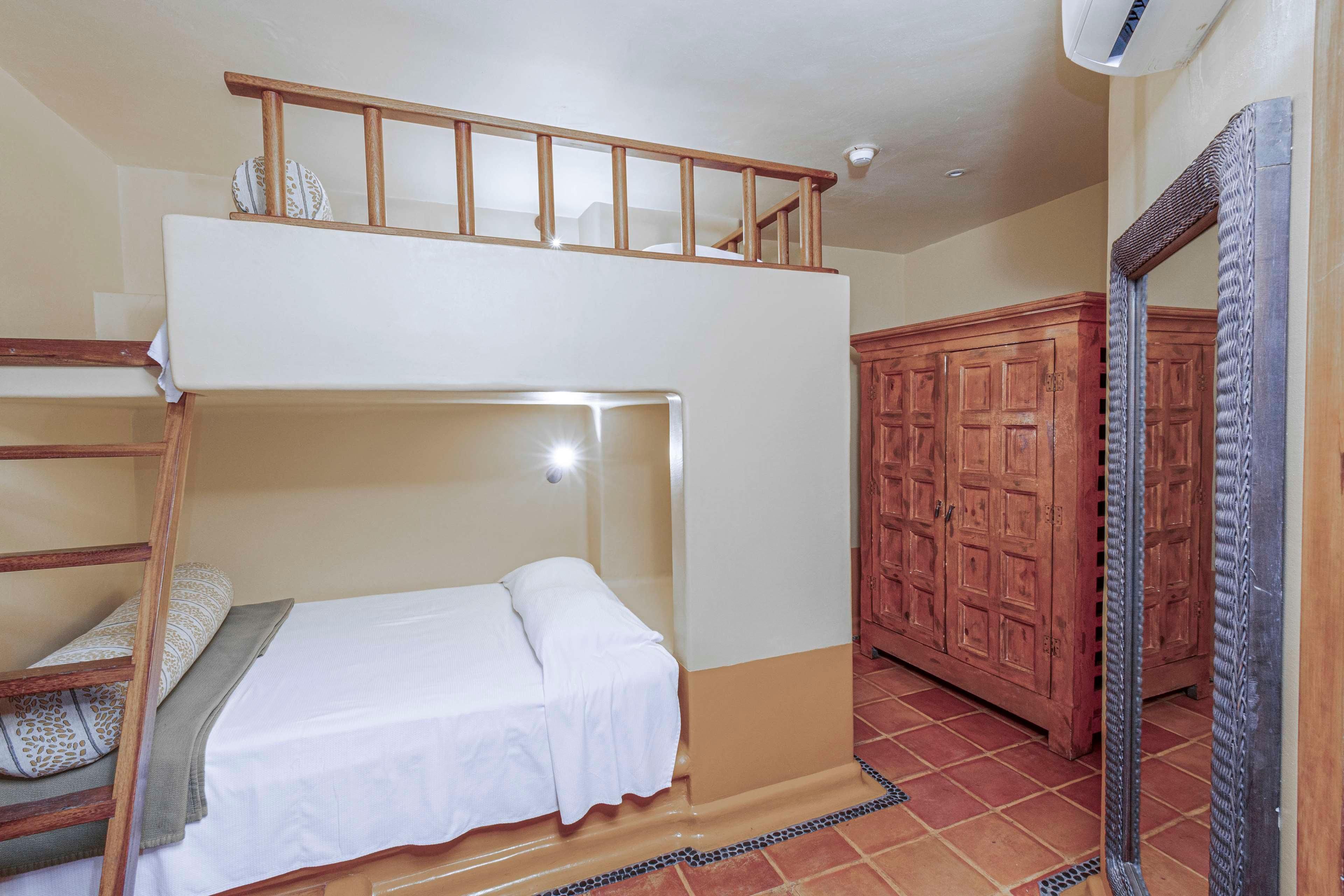 Una cama litera con escalera de madera, una cama individual con sábana blanca, un armario y un espejo.