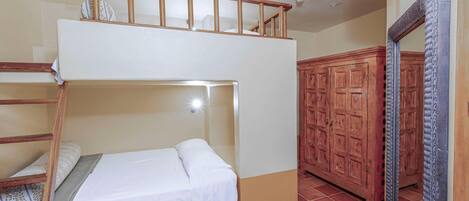 Una cama litera con escalera de madera, una cama individual con sábana blanca, un armario y un espejo.