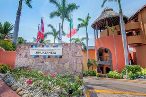Señal del Hilton Grand Vacations Club Zihuatanejo, con palmeras y una estructura de techo de paja al fondo.