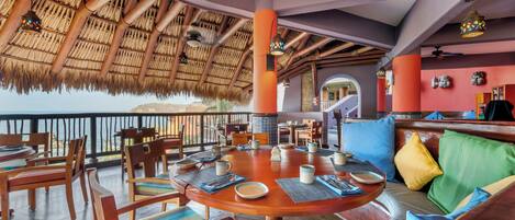 Restaurante com telhado de palha, móveis de madeira e vista para o mar.