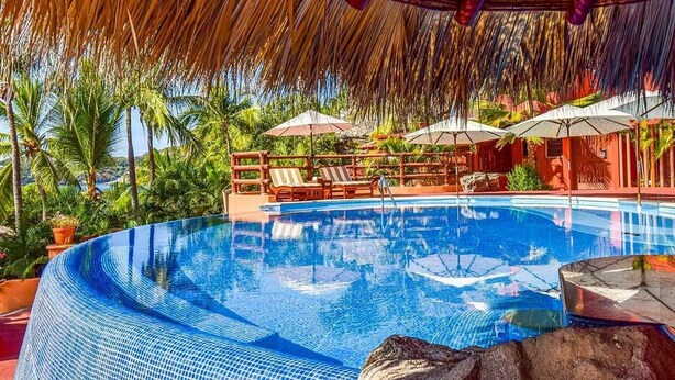 A clear blue infinity pool with lounge chairs and umbrellas, surrounded by lush greenery and a thatched roof structure. - Hilton Grand Vacations Club Zihuatanejo (Zihuatanejo)