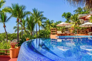 A clear blue infinity pool with lounge chairs and umbrellas, surrounded by palm trees and a stone wall. - Hilton Grand Vacations Club Zihuatanejo (Zihuatanejo)