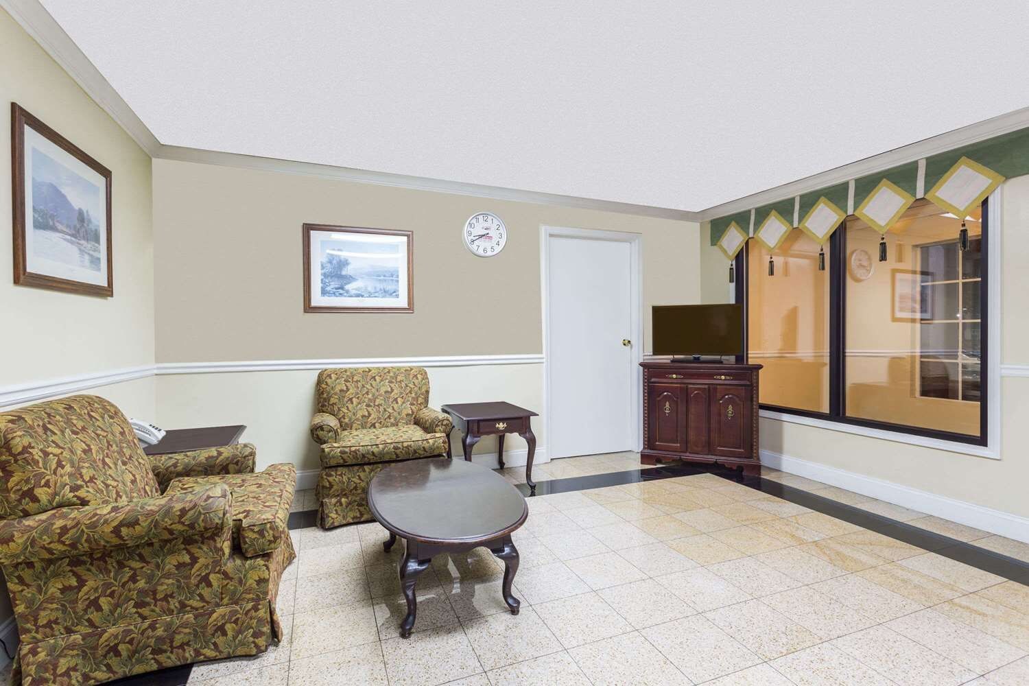 A room with two patterned armchairs, a small table, a clock on the wall, a television, and a cabinet with a mirror.