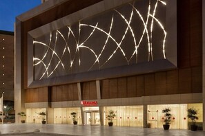 Un bâtiment moderne avec une grande installation lumineuse illuminant la façade, une enseigne indiquant « RAMADA » et des plantes en pot devant l’entrée.