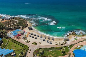 Vue aérienne d’un hôtel situé sur la côte, avec une plage, une piscine et un parking.