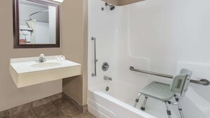 Un baño con un lavabo blanco, un espejo, una ducha con barra de apoyo y un asiento de ducha accesible para sillas de ruedas.