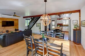 A modern living room with a wooden floor, a black leather sofa, a dining table with chairs, and a staircase leading to the upper floor.