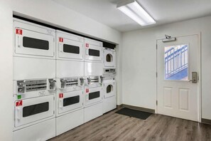 A laundry room with multiple white machines, numbered from 6 to 11, and a door with a view of a building outside. - Suburban Studios Lakeland North I-4 (Lakeland)