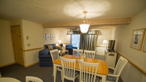 A dining area with a wooden table and white chairs, a sofa, a lamp, and a television on a stand.