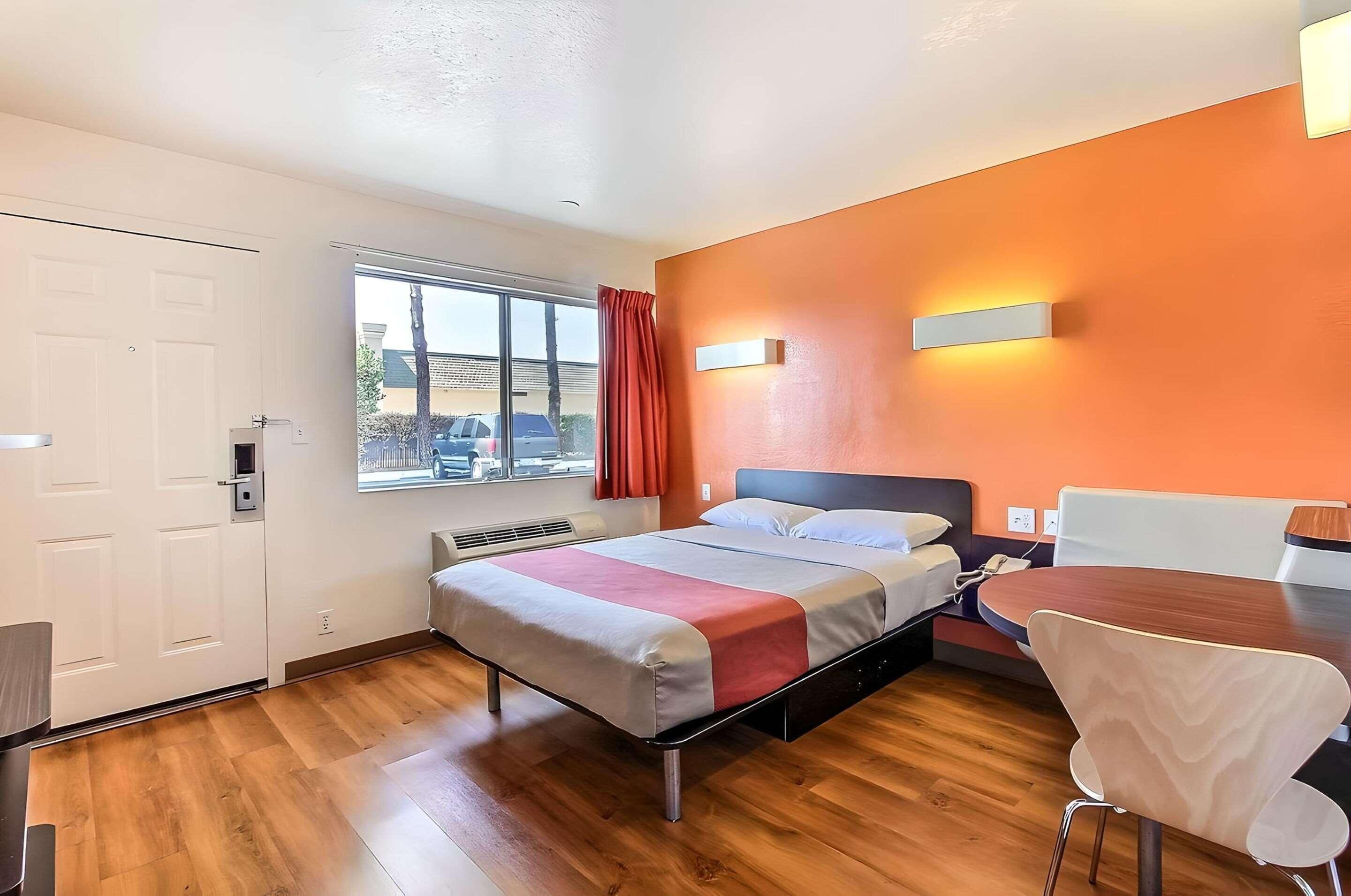 A hotel room with a bed, a desk, a chair, a window with curtains, and an orange wall.