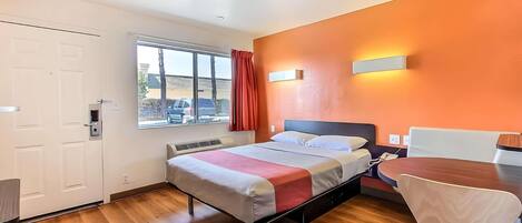 A hotel room with a bed, a desk, a chair, a window with curtains, and an orange wall.