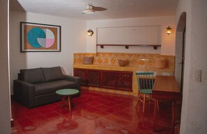 Ein kompakter Wohnraum mit einer Couch, einem kleinen runden Tisch, einem Esstisch mit Stühlen, einer Küchenzeile mit einer Arbeitsplatte und Barhocker sowie einem Deckenventilator.