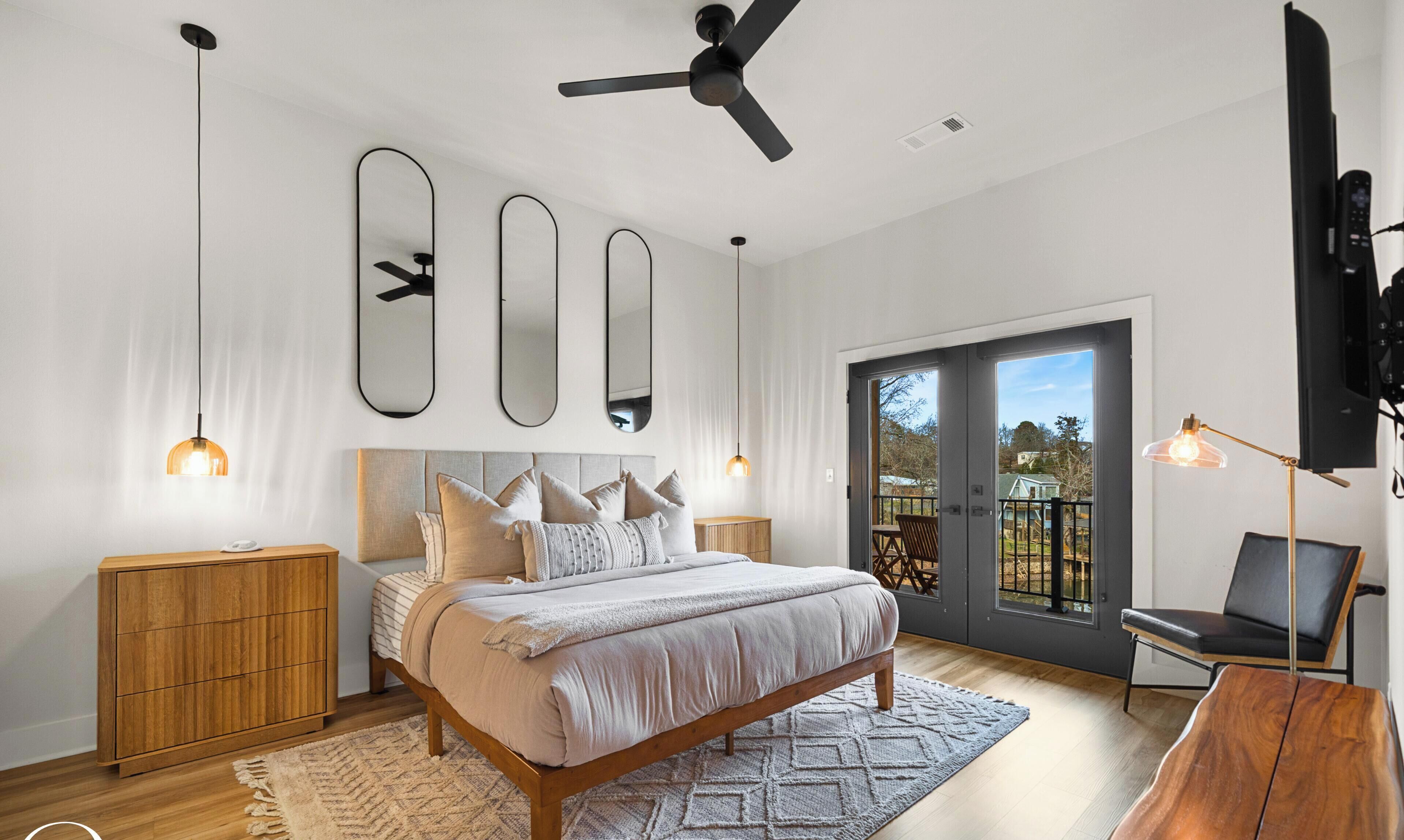 Une chambre à coucher comprenant un lit, des tables de chevet, un ventilateur de plafond, un grand miroir et une vue sur l’extérieur, accessible par une porte-fenêtre coulissante.