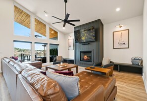 A spacious living room with a leather sofa, a fireplace, a television mounted on the wall, and large windows offering a view of the outdoors. - The Great Escape - 1 (Hot Springs)