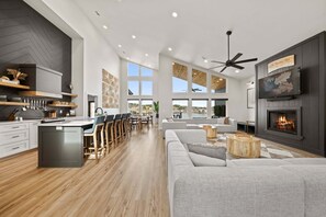 Eine moderne Küche mit Kochinsel, Essbereich mit Stühlen, ein graues Sofa, ein Kamin und große Fenster.