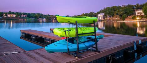 A wooden dock with kayaks and a paddleboard, overlooking a calm lake with houses in the background.