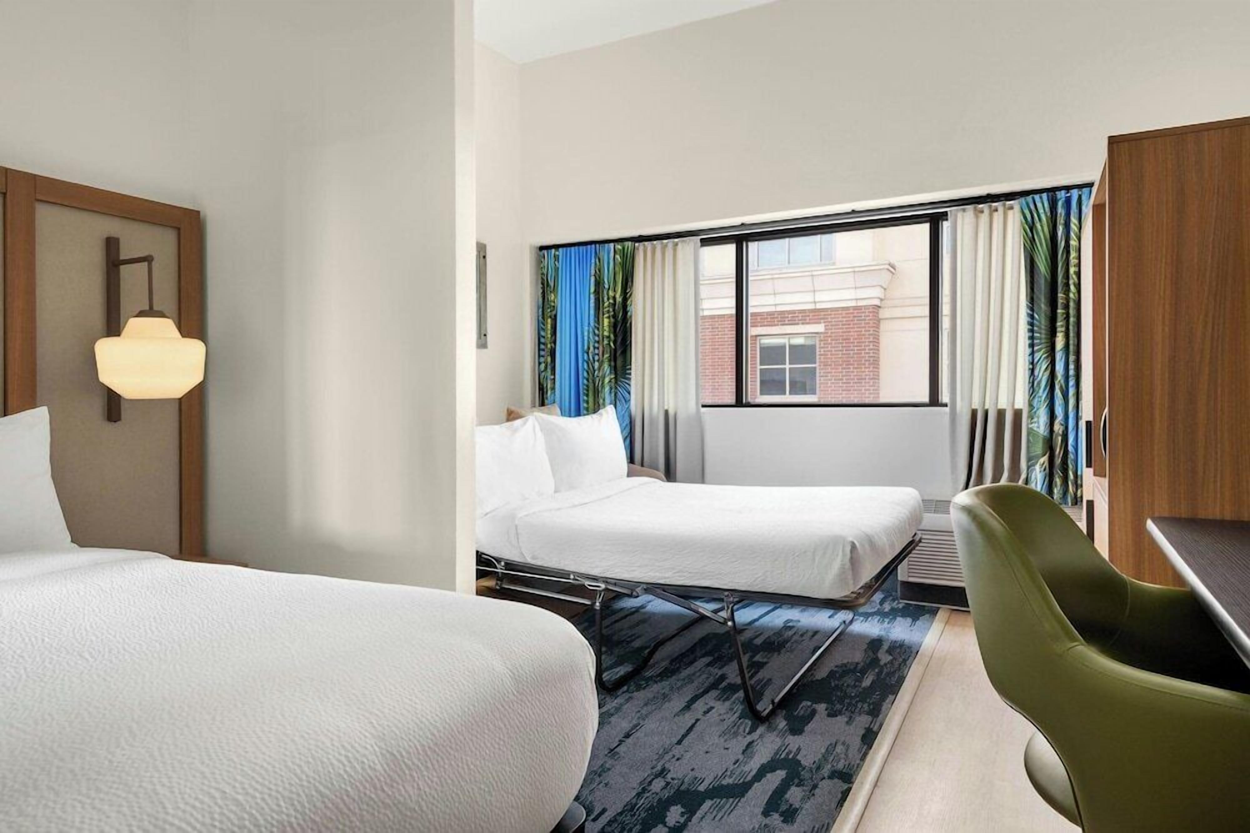 Une chambre d’hôtel avec deux lits, un bureau, une chaise et une grande fenêtre avec des rideaux.
