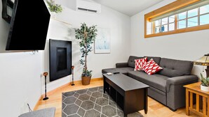A living room with a flat-screen TV, a ceiling fan, a large potted plant, a grey sofa with red pillows, and a wooden side table.