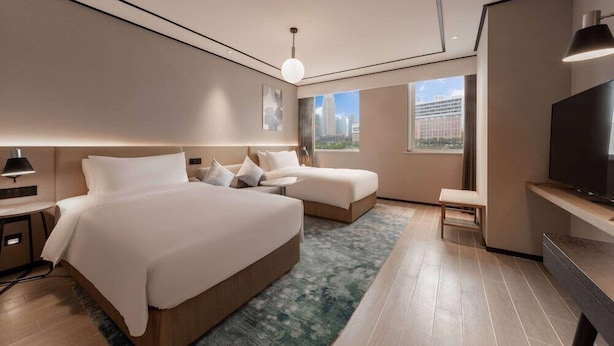 Ein modernes Hotelzimmer mit zwei Betten, einem Fernseher, einem Schreibtisch und einem großen Fenster mit Blick auf die Stadt.