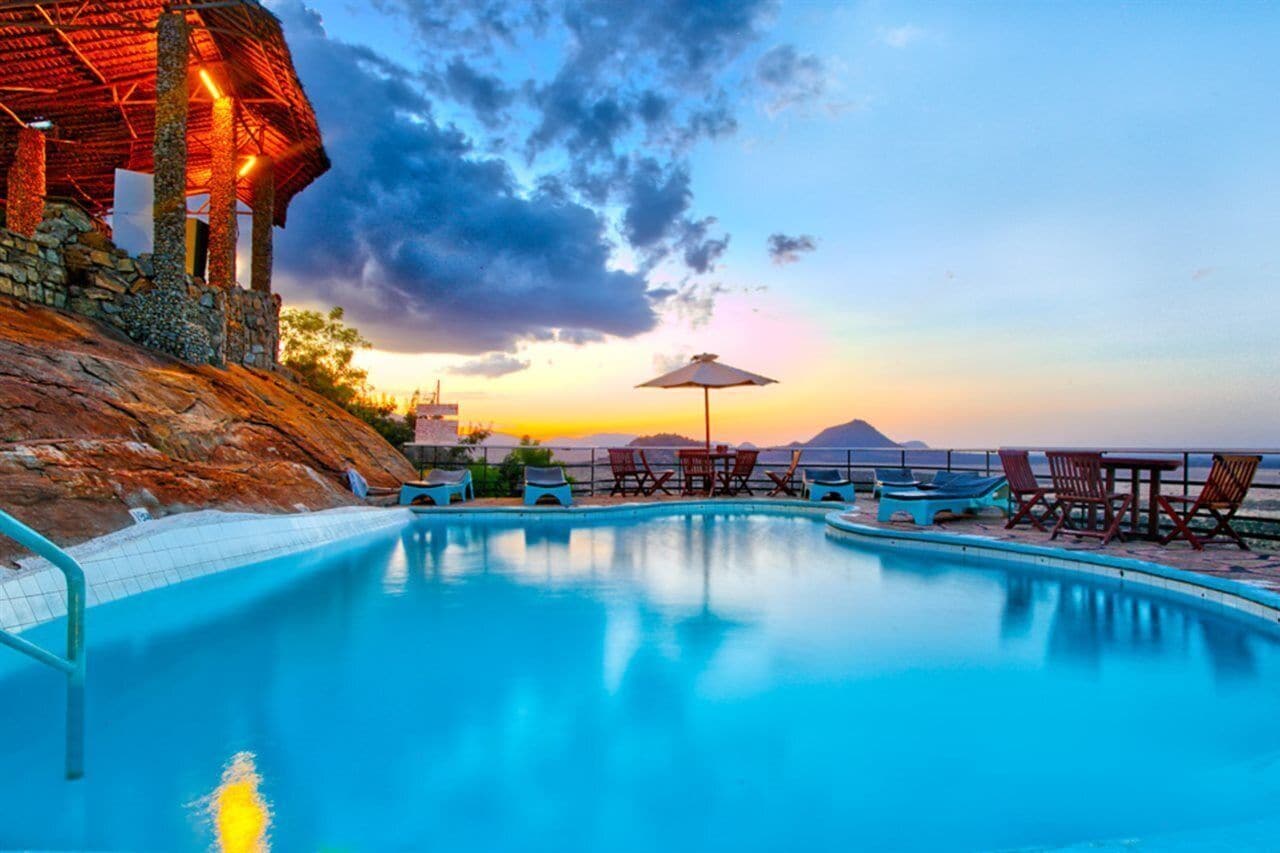 A swimming pool with lounge chairs and a table, surrounded by stone walls and a view of a sunset over mountains.