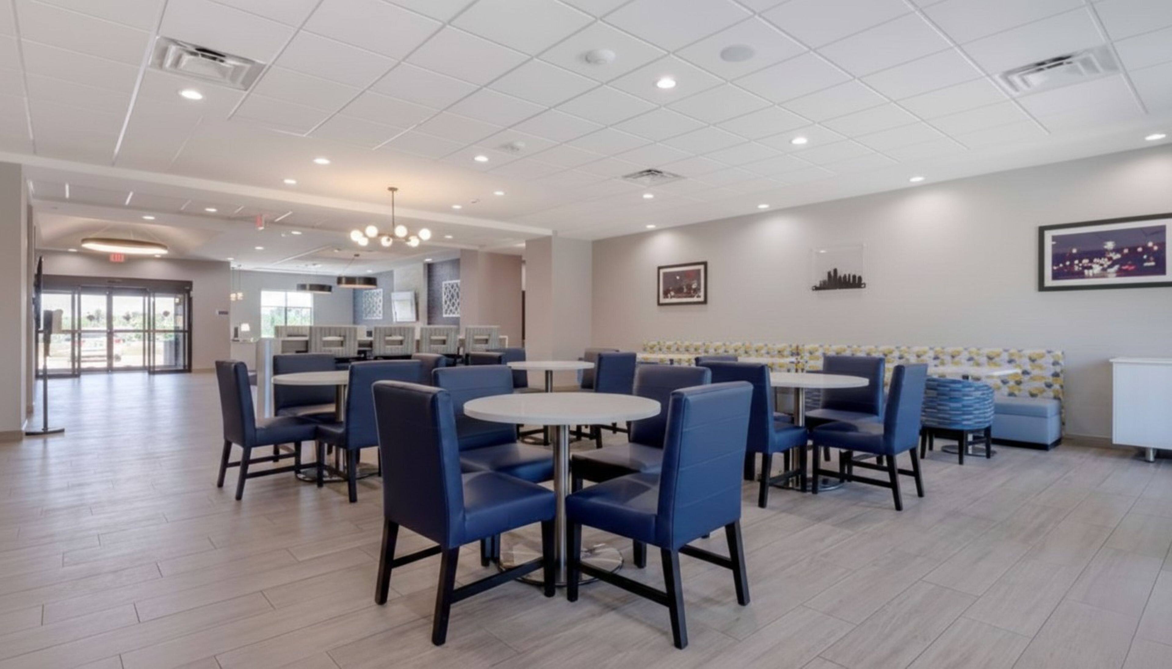 A modern dining area with round tables and blue chairs, a kitchen in the background, and a cityscape artwork on the wall.