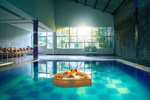 An indoor swimming pool with a sunlit view, a lounge area with chairs, and a decorative centerpiece on the pool. - THE Fern Surya Resort (Kasauli)