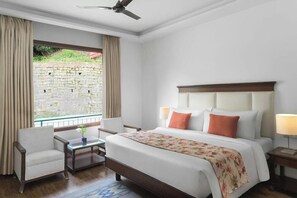 A hotel room with a bed, two bedside tables, a chair, a small table with a vase, a ceiling fan, and a view of a stone wall and greenery outside. - THE Fern Surya Resort (Kasauli)