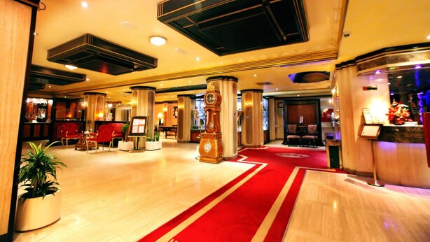 A hotel lobby with a red carpet, wooden columns, a reception desk, and a clock.