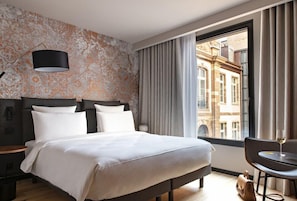 A hotel room with a large bed, a nightstand, a lamp, a window with curtains, and a view of buildings outside. - Leonor Hotel (Strasbourg)