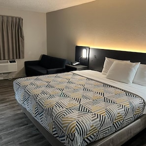 A hotel room with a bed, a desk, a chair, and a wall-mounted lamp. - Motel6 Tulsa, OK - Airport (Tulsa)