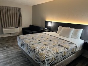 A hotel room with a bed, a chair, a bedside table with a lamp, and an air conditioning unit.