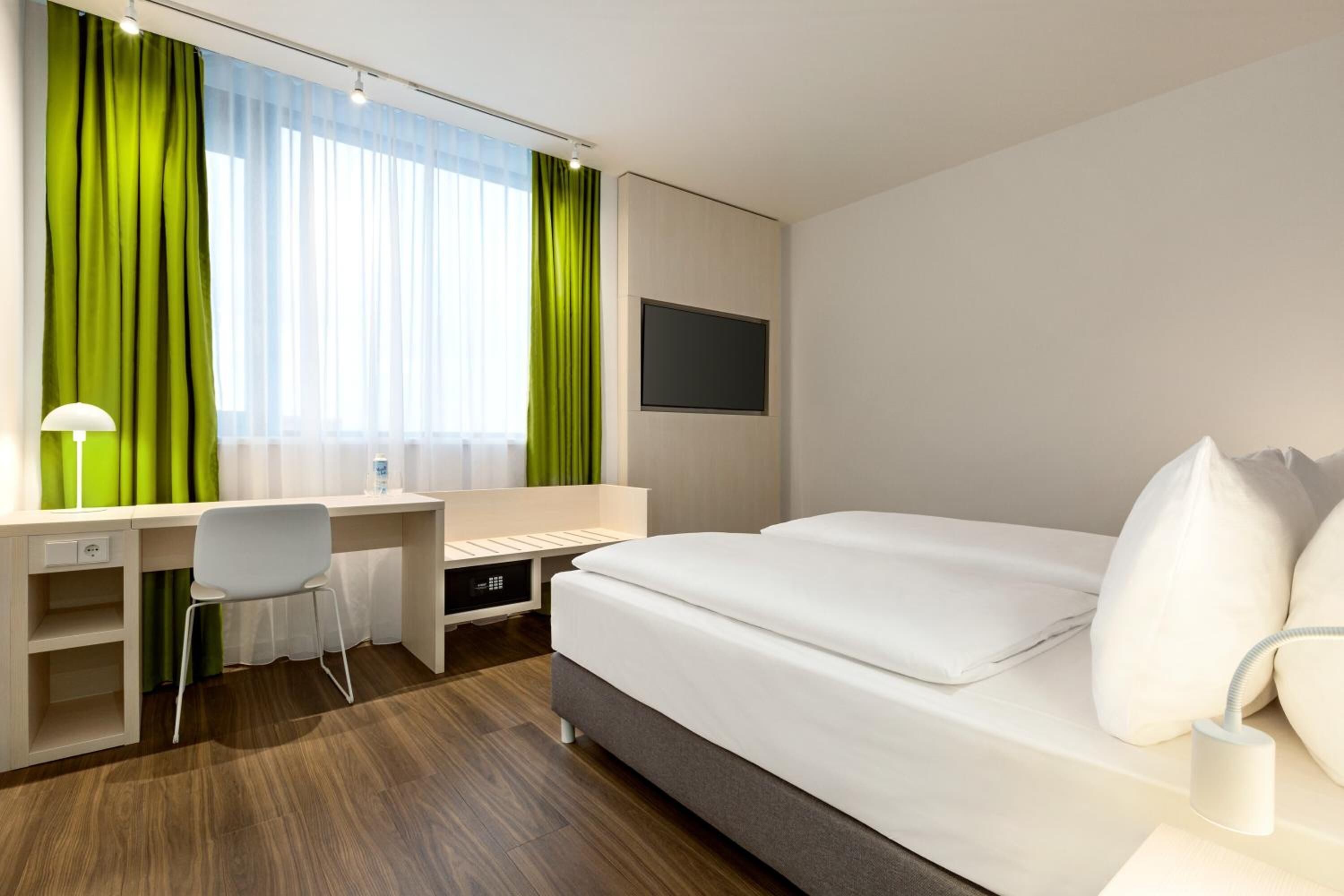 Ein modernes Hotelzimmer mit Bett, Schreibtisch, Stuhl und einem an der Wand befestigten Fernseher.