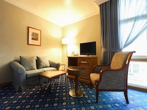Una camera d'albergo con un divano, una poltrona, televisione, una lampada e un tappeto decorato.